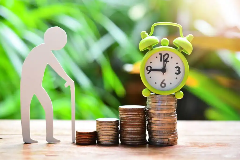 A paper cutout of an elderly person with a cane stands beside stacked coins and a green alarm clock, symbolizing saving for retirement. The background is blurred greenery.
