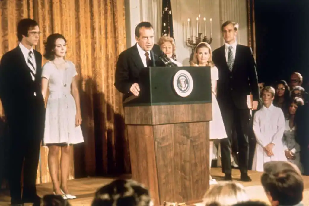 President Richard Nixon speaks at a podium with the presidential seal, flanked by his family and others in formal attire, in front of golden curtains and an American flag, during a public event.