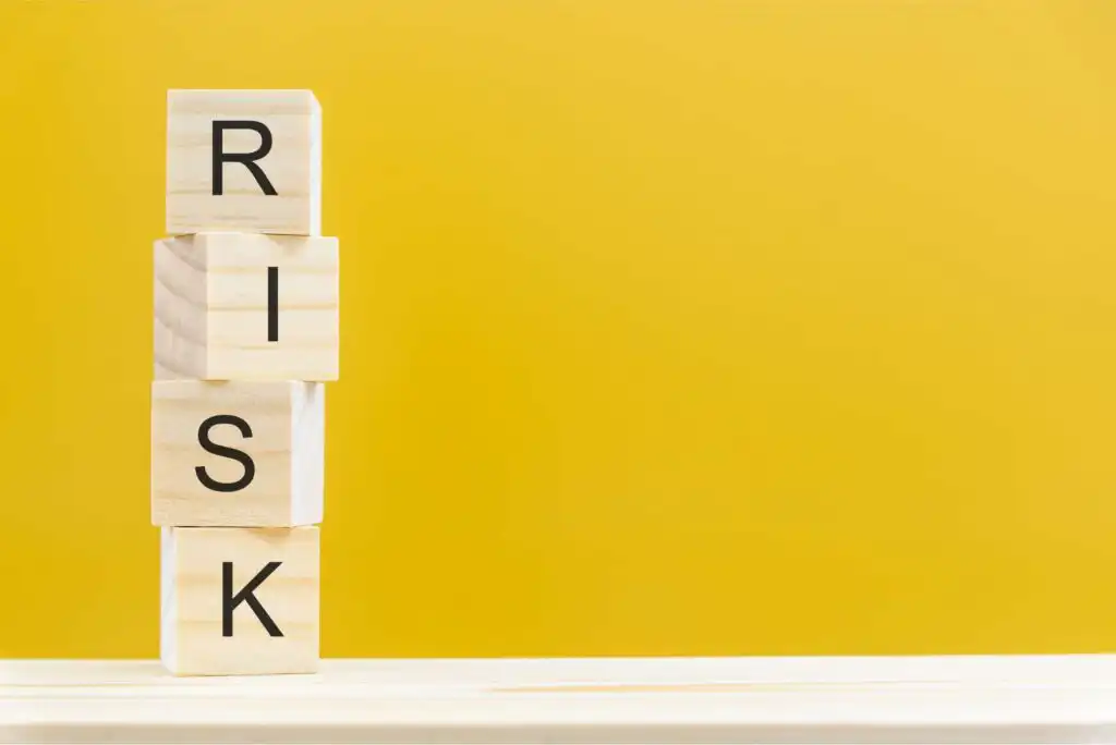 Four wooden blocks stacked vertically spell RISK against a plain yellow background, with empty space to the right.