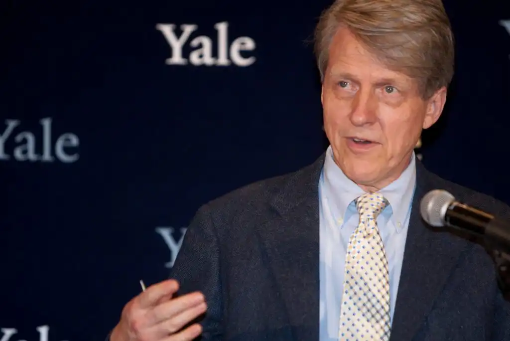 A man in a suit and polka dot tie speaks at a microphone in front of a dark blue backdrop with the word Yale repeated in white letters.