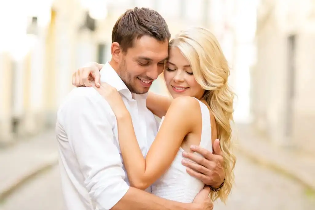 A smiling couple embraces affectionately outdoors, both wearing white shirts. The woman has long blonde hair and closed eyes, while the man, with short brown hair, gently holds her. The background is softly blurred.