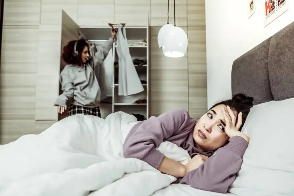 A woman lies in bed looking stressed while another woman stands by an open wardrobe in the background, holding up a dress as if deciding what to wear. The scene is set in a modern bedroom.