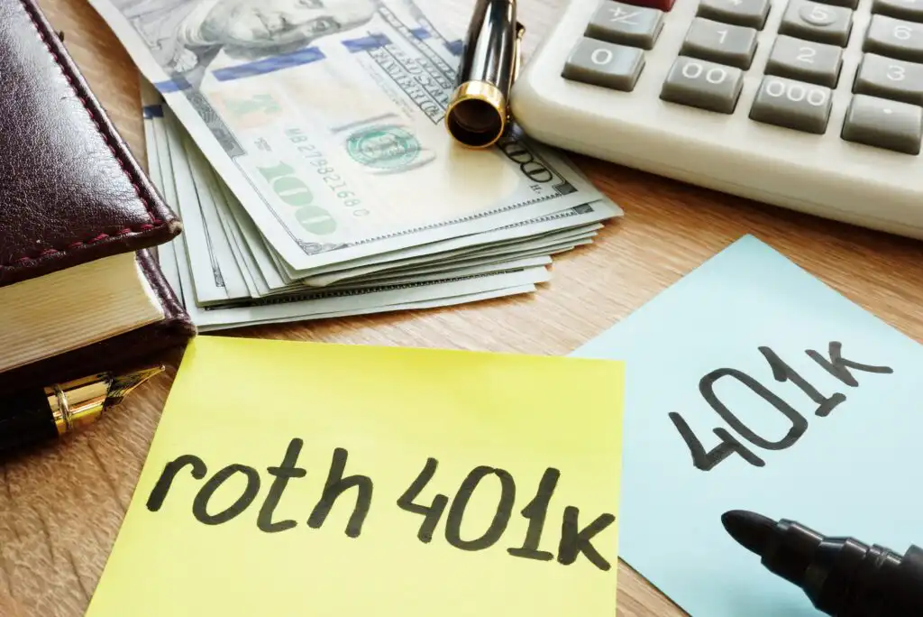 A desk with a stack of $100 bills, a pen, a calculator, and sticky notes labeled “roth 401k” and “401k,” highlighting a comparison of retirement savings options.