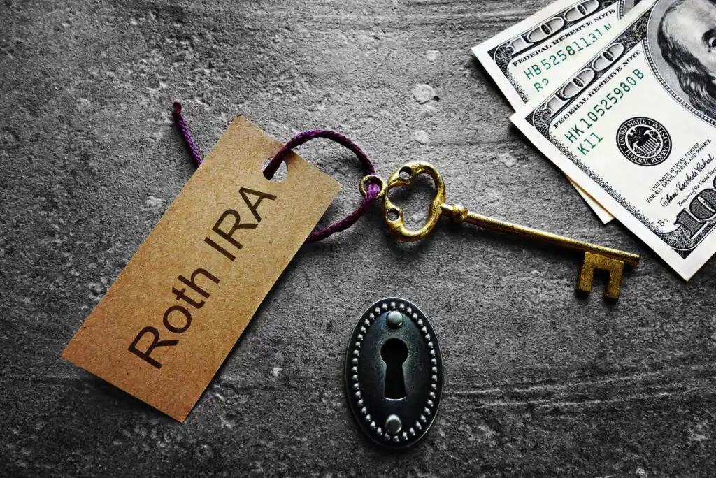 A key with a tag labeled Roth IRA lies next to a keyhole and several one hundred dollar bills on a dark textured surface.