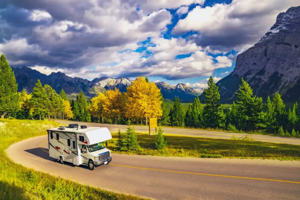A white RV drives along a winding road bordered by green trees and autumn foliage, with tall mountains and dramatic clouds in the background under a vibrant blue sky.