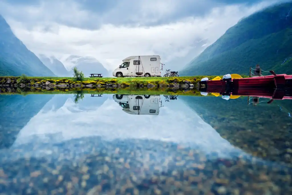 A camper van is parked beside a calm mountain lake, with its reflection mirrored in the water. Nearby are a picnic table, two bicycles, and red boats, surrounded by lush green mountains under a cloudy sky.