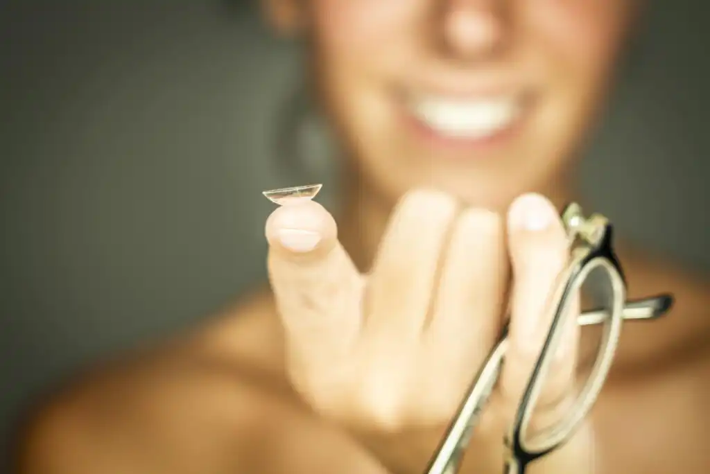 A person smiling holds a contact lens on their index finger, with a pair of eyeglasses in their other hand. The background is blurred, focusing on the contact lens.