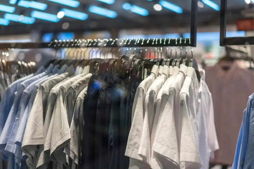 A row of shirts in various colors, including white, blue, and gray, hanging on black hangers on a clothing rack in a store, with blurred lights and shelves in the background.