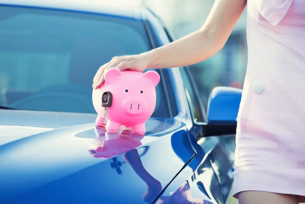 A person in a light pink outfit places a hand on a pink piggy bank with a car key on top, resting on the hood of a shiny blue car.
