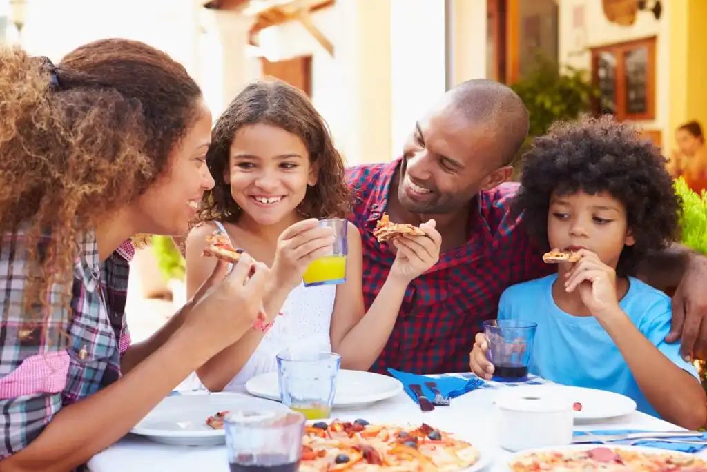 A family of four sits at an outdoor table, smiling and eating pizza together. The parents and two children enjoy their meal, with drinks and pizza visible on the table.