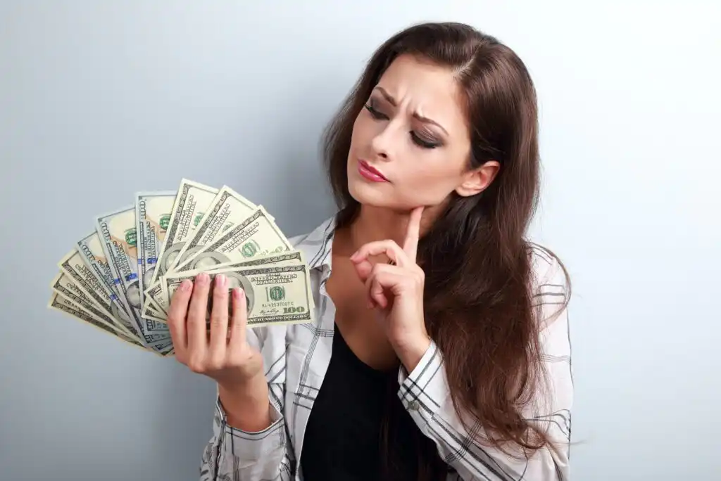 A woman with long brown hair holds a fan of U.S. dollar bills in one hand and thoughtfully touches her chin with the other, looking at the money with a questioning expression.