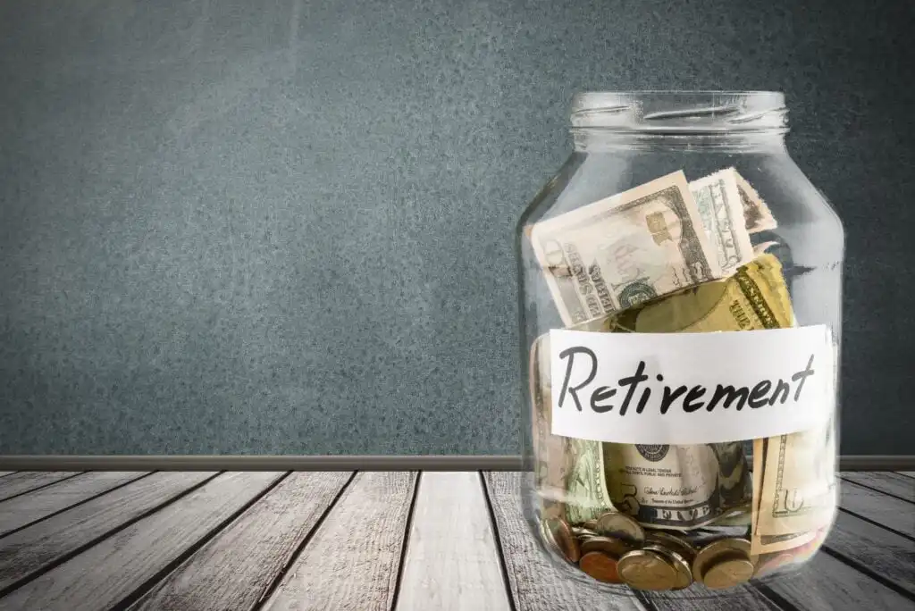 A glass jar labeled Retirement filled with U.S. dollar bills and coins sits on a wooden surface against a chalkboard background.