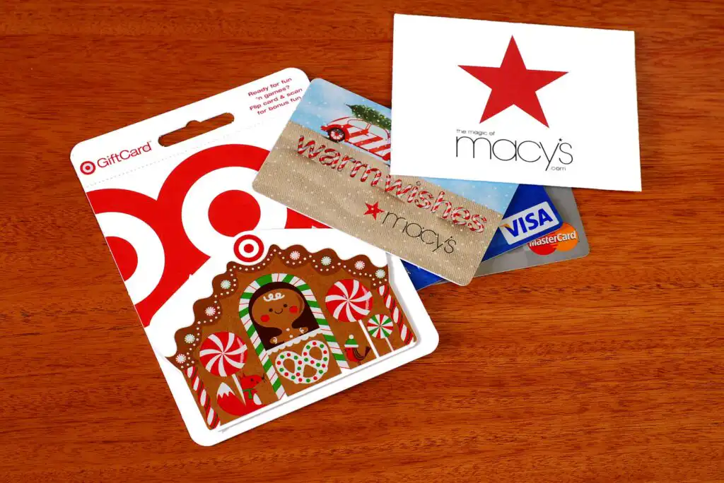 Gift cards from Target, Macys, and Visa are displayed on a wooden surface. The Target card features a gingerbread house with candy decorations, while the Macys card says warm wishes with a red star logo.