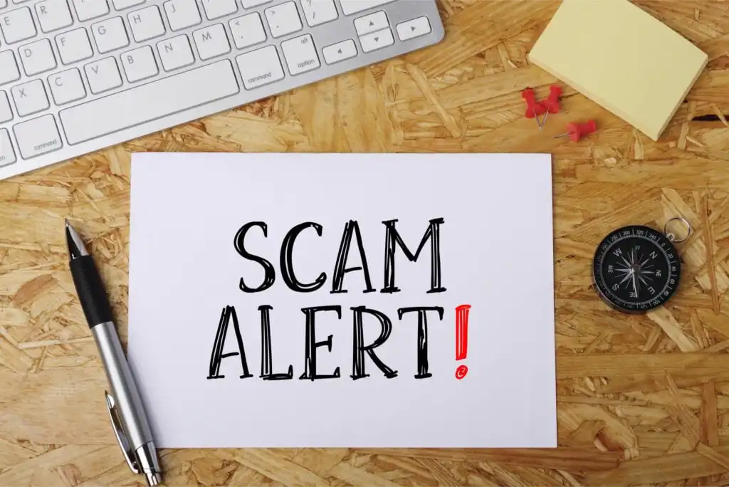 A white paper with the words SCAM ALERT! in bold letters lies on a wooden desk next to a pen, compass, keyboard, yellow sticky notes, and red push pins.