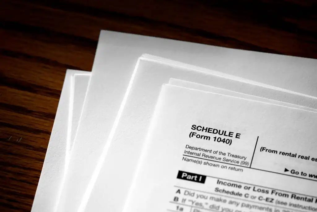 A close-up of several stacked tax documents on a wooden surface, with the top page showing part of a Schedule E (Form 1040) from the IRS.