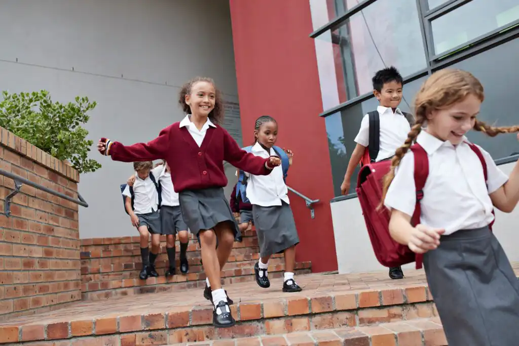 A group of smiling schoolchildren in uniforms walk and run down brick steps outside a school building, carrying backpacks and looking happy.