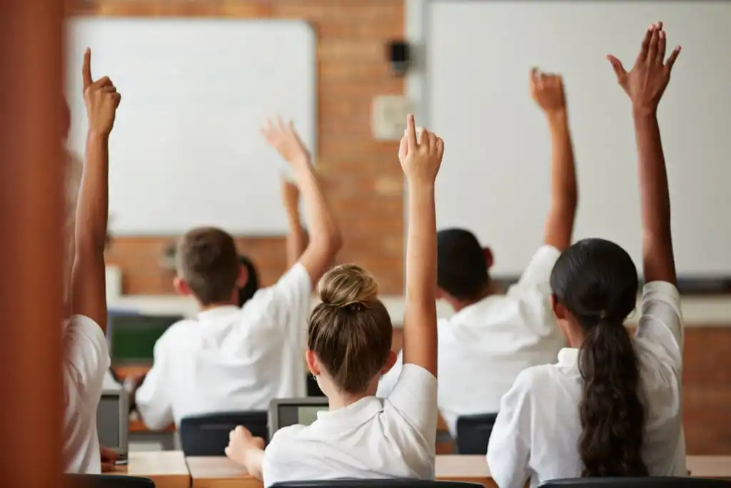 Several students in white uniforms sit at desks in a classroom with their hands raised, facing a whiteboard at the front of the room. Their backs are to the camera.