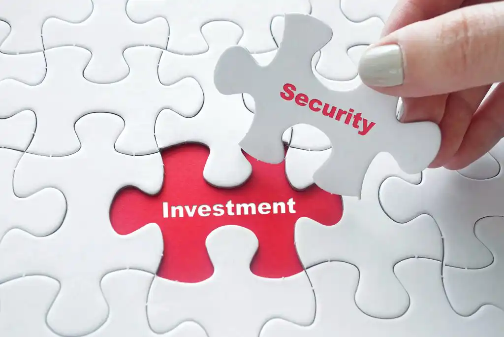 A hand places a puzzle piece labeled “Security” into a puzzle with a missing piece, revealing the word “Investment” underneath on a red background.