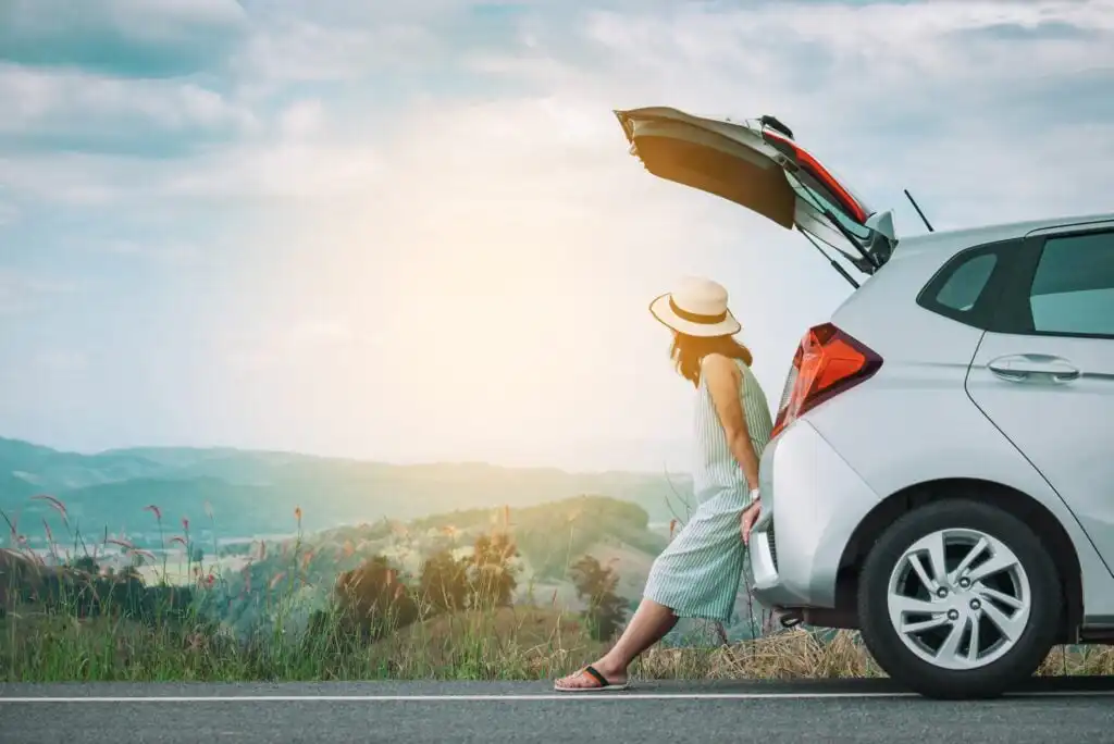 A woman in a dress and hat sits on the back of a silver car with its trunk open, parked by the roadside. She faces a scenic landscape of hills and sky during daylight.