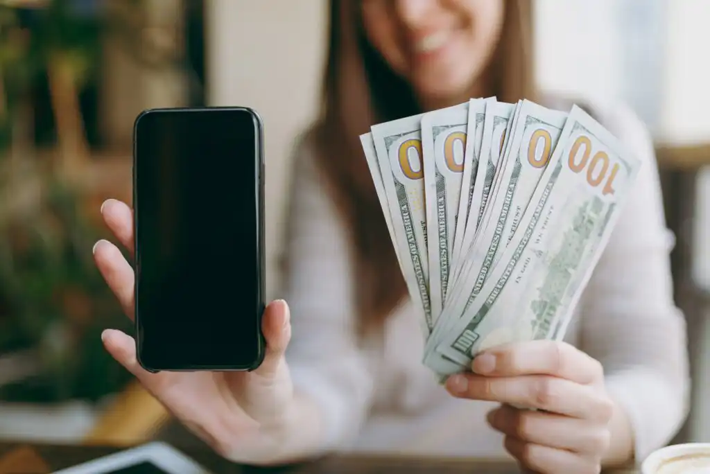 A woman holds a blank smartphone in one hand and several $100 bills in the other, smiling in the background. The focus is on the money and phone.