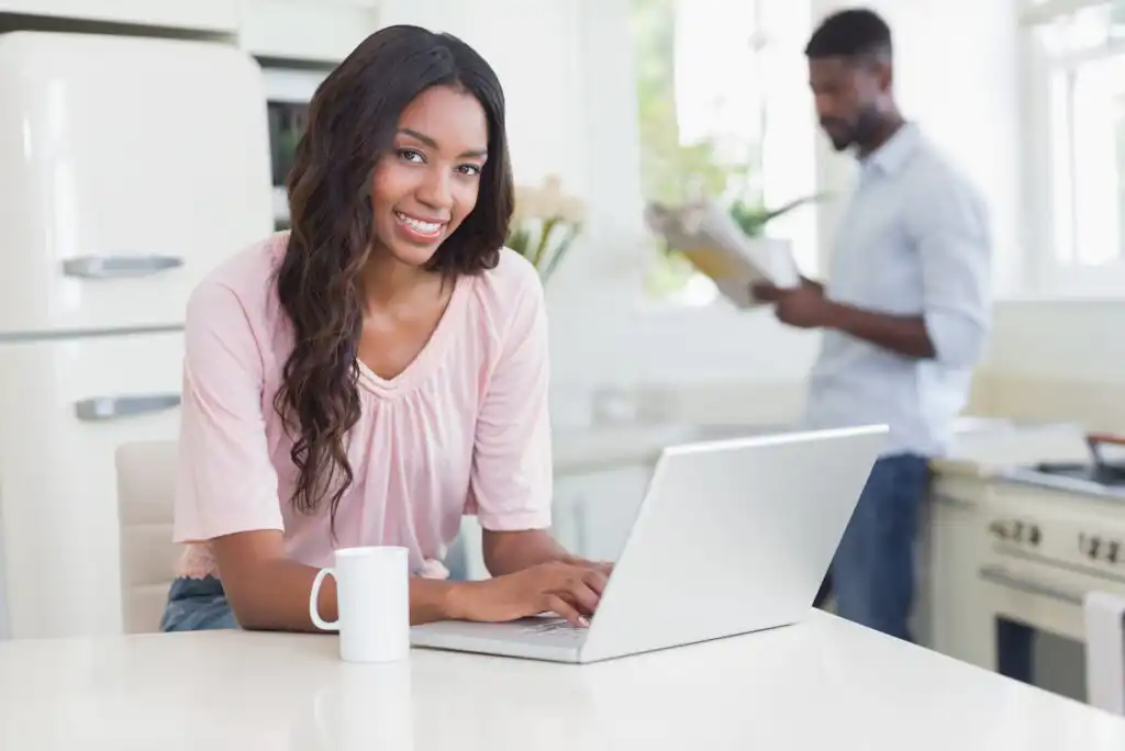 A woman smiles while using a laptop at a kitchen counter with a white mug nearby. In the background, a man stands by the stove reading a newspaper. The setting is bright and modern.
