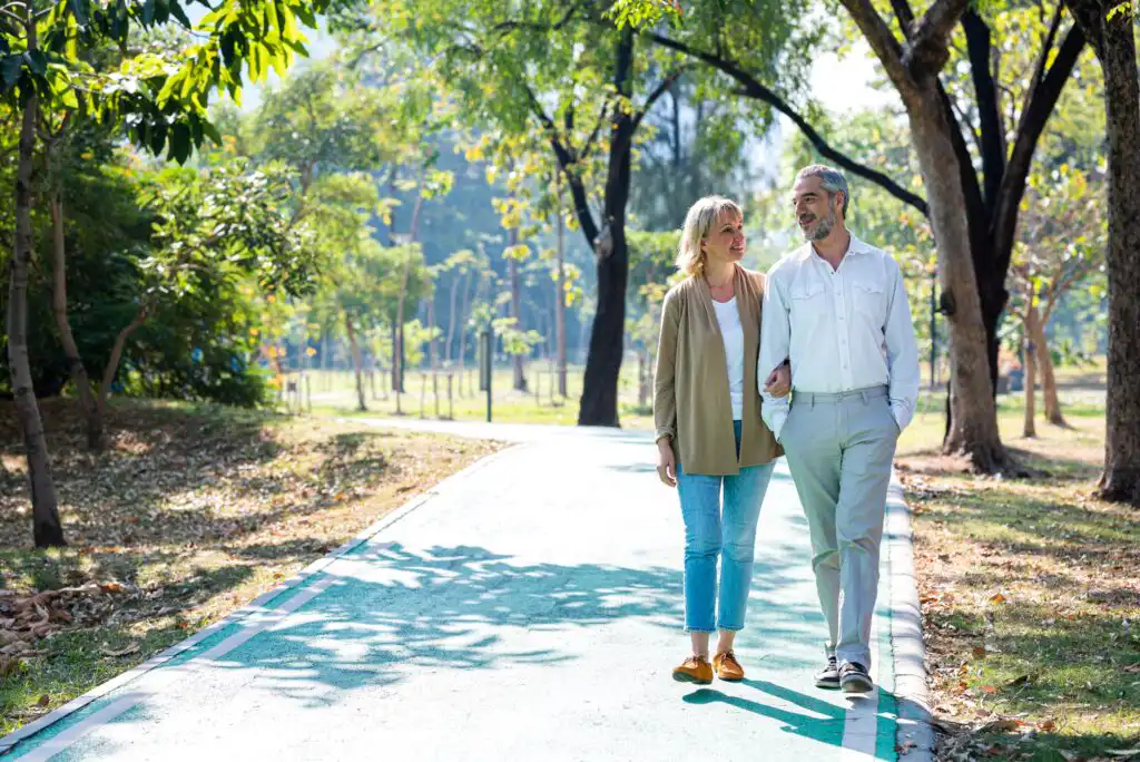 An older couple walks arm in arm along a sunlit path in a green, leafy park, smiling and enjoying each others company on a pleasant day.