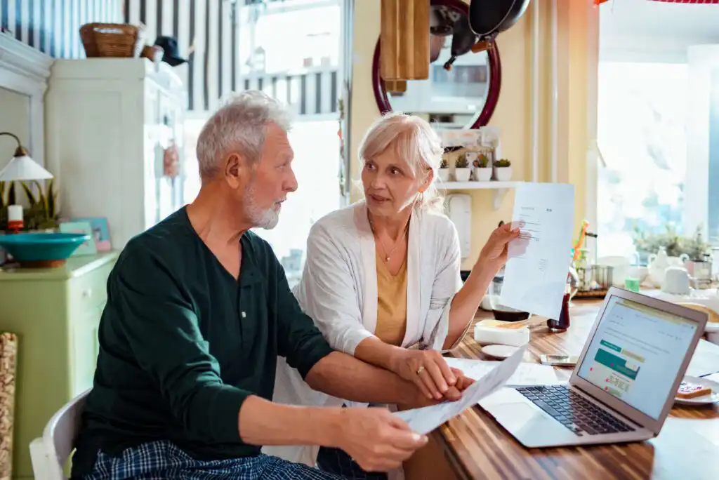 An older couple sits at a kitchen table, discussing paperwork. The woman holds a document while the man looks at her, and an open laptop sits on the table next to them. They appear to be reviewing finances together.
