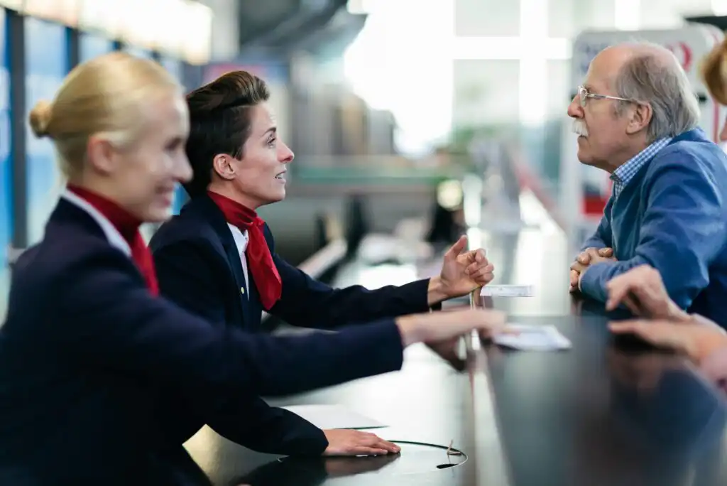 Two airline staff in uniform assist an older man at an airport check-in counter. The staff are smiling and holding documents while the man, leaning on the counter, listens attentively. The background is softly blurred.