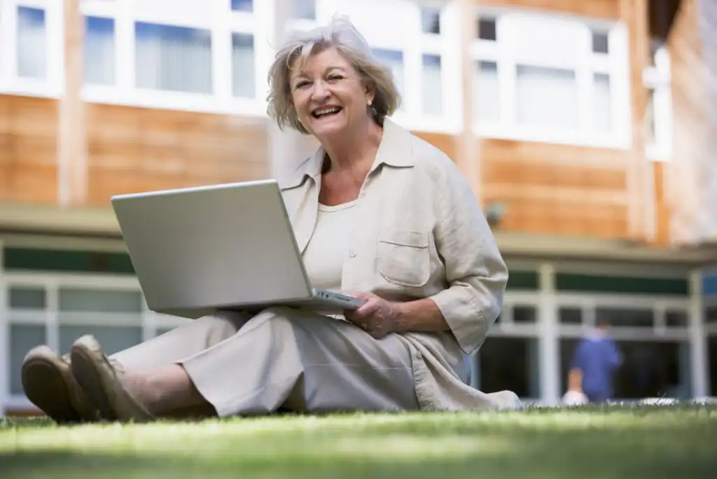 An older woman sits on grass outside a building, smiling while using a laptop on her lap. She wears a light-colored outfit and appears to be enjoying her time working or browsing outdoors.