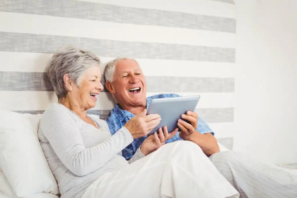 An elderly couple sits on a bed, smiling and laughing together while looking at a tablet. They appear relaxed and joyful in a cozy, striped-bedroom setting.