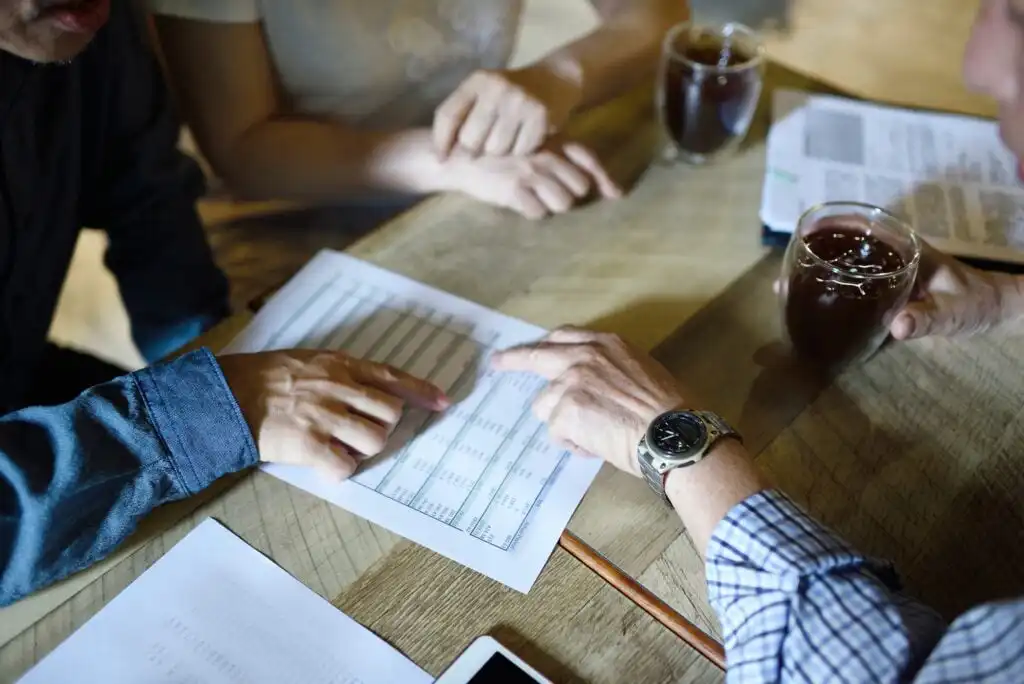 Three people sit around a wooden table, discussing and pointing at a printed spreadsheet. Two cups of coffee, documents, and a smartphone are also on the table. Only their hands and arms are visible.