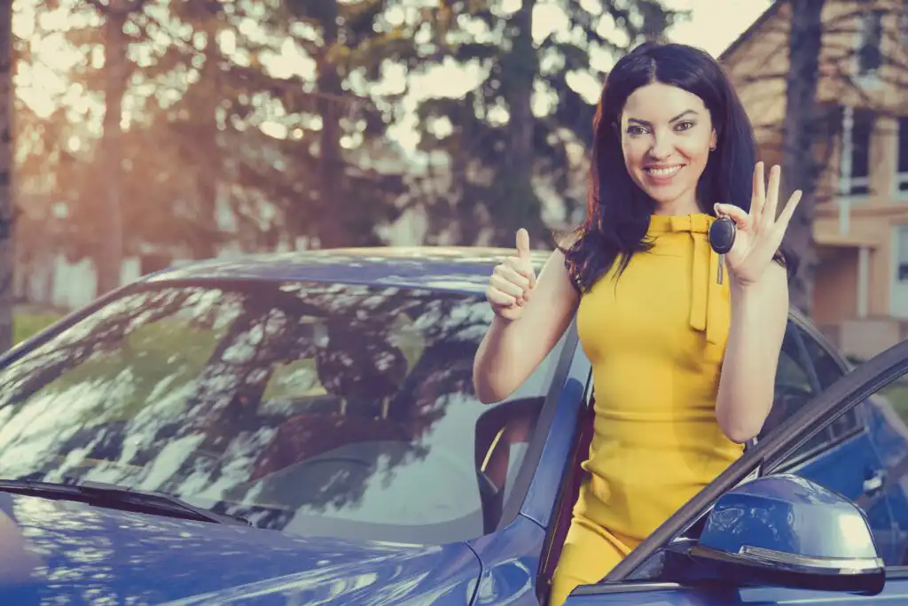 A smiling woman in a yellow dress stands by a blue car, holding a car key in one hand and giving a thumbs-up with the other, suggesting she is happy and possibly just bought or received the car.