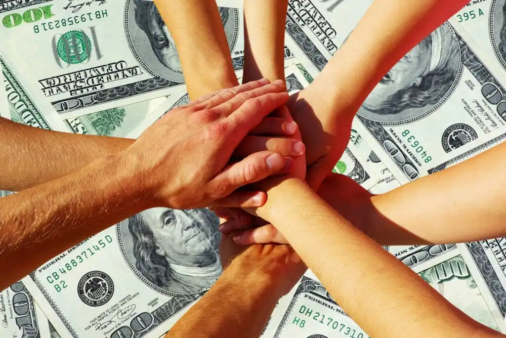 Several hands stacked together in a gesture of unity, teamwork, or agreement, with a background of multiple hundred-dollar bills spread out underneath.
