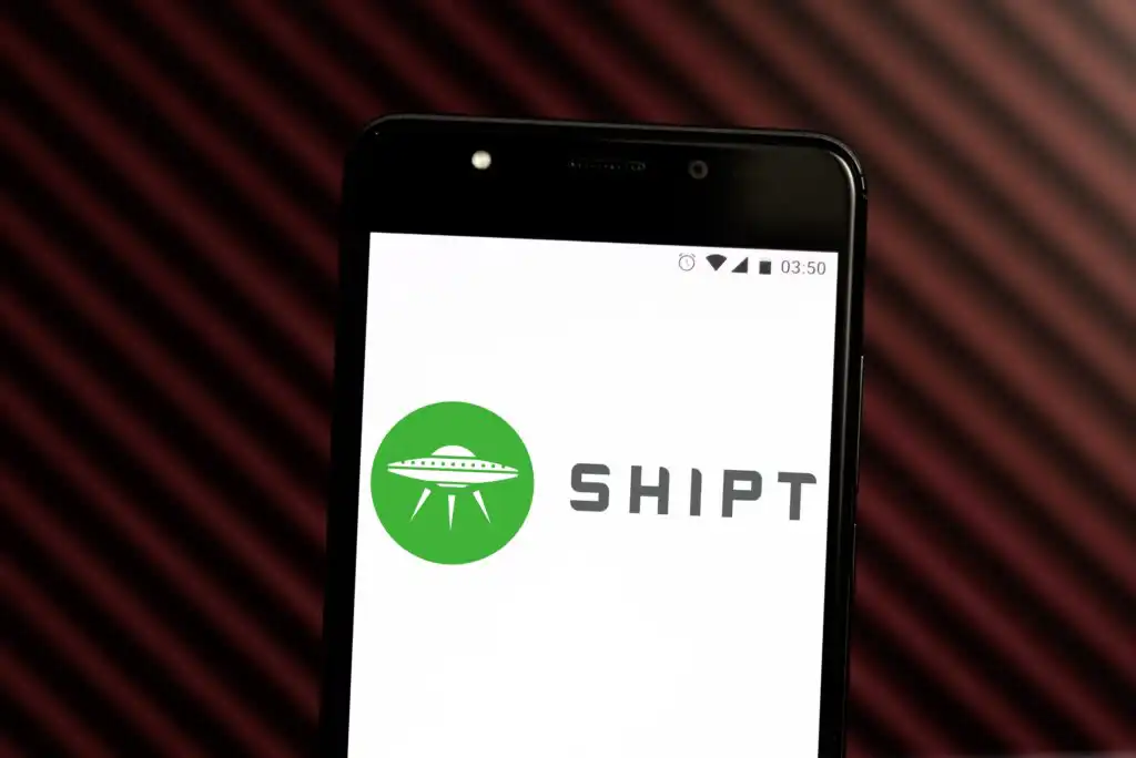 A smartphone screen displaying the Shipt logo, featuring a green circle with a white UFO, against a background with diagonal maroon and black stripes.