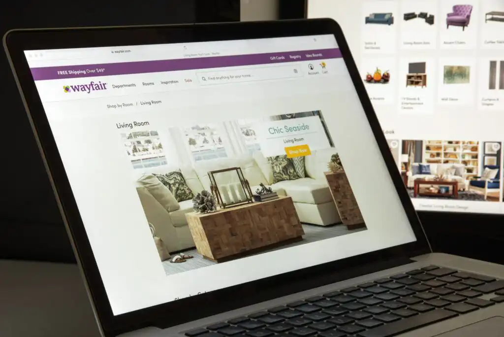 A laptop displaying the Wayfair website’s living room furniture section; the screen shows a featured image of a stylish living room with the header “Chic Seaside” and various decor items.