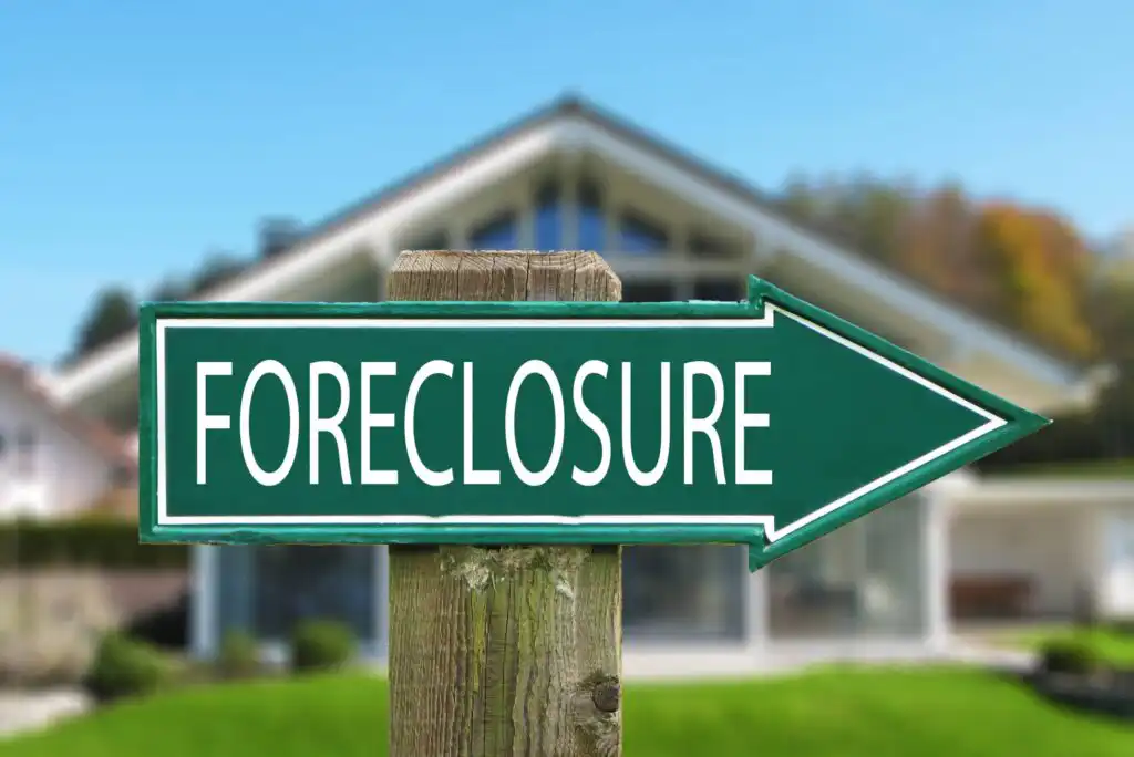 A green arrow-shaped sign with the word Foreclosure points toward a house in the background, which appears out of focus.