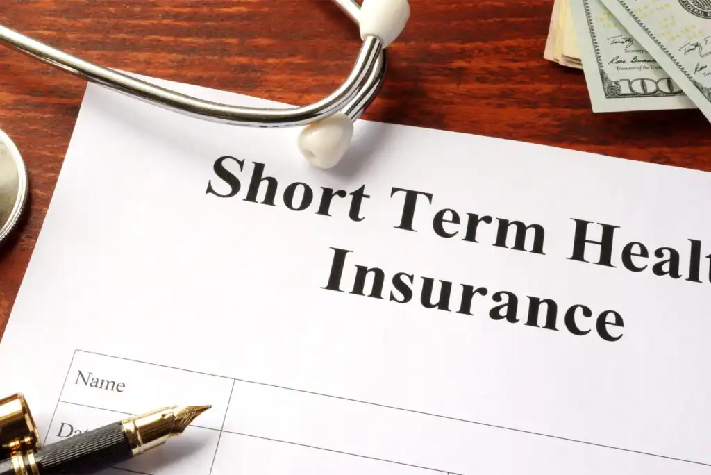 A document titled Short Term Health Insurance is on a desk, with a stethoscope, fountain pen, and a stack of cash nearby, suggesting a focus on healthcare and financial planning.