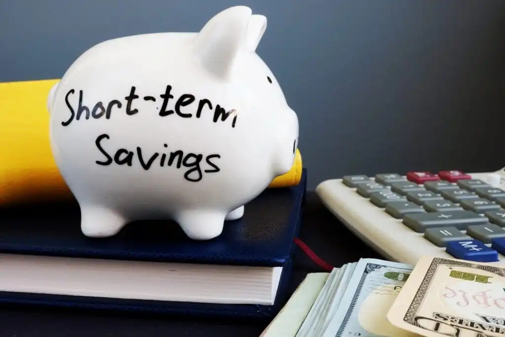 A white piggy bank labeled Short-term Savings sits on a stack of books next to a calculator, cash, and a credit card, symbolizing personal finance and saving money.