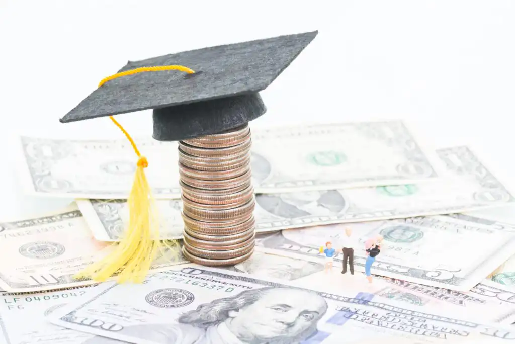 A stack of coins topped with a graduation cap sits on scattered U.S. dollar bills, with small figurines of adults and a child standing nearby, symbolizing education costs and student loans.