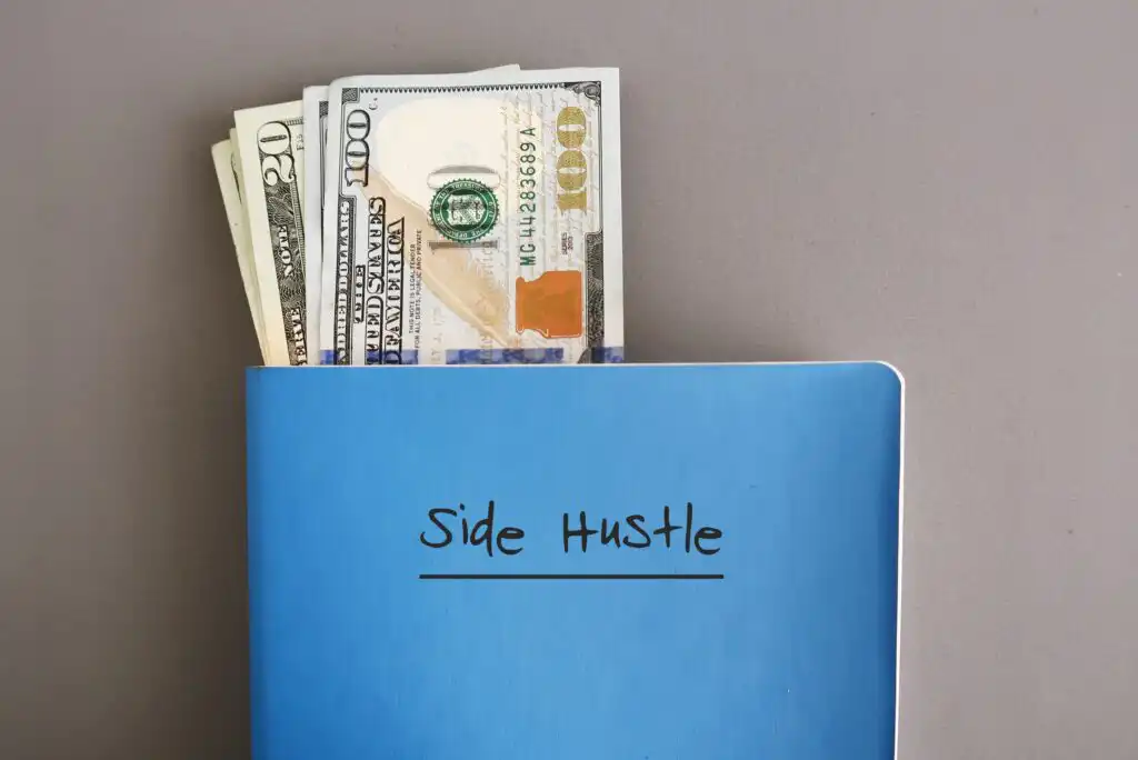A blue notebook labeled Side Hustle with several U.S. dollar bills, including $20 and $100 notes, sticking out from the top, placed on a gray surface.