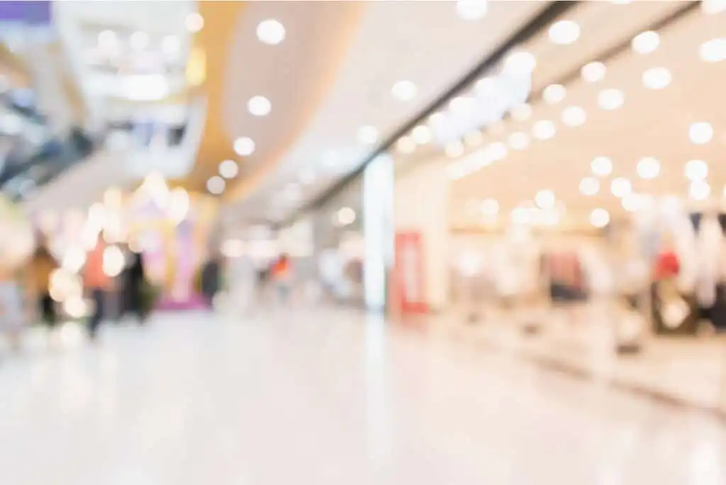A blurred image of the interior of a shopping mall with bright lights, storefronts, and indistinct figures walking in the background.