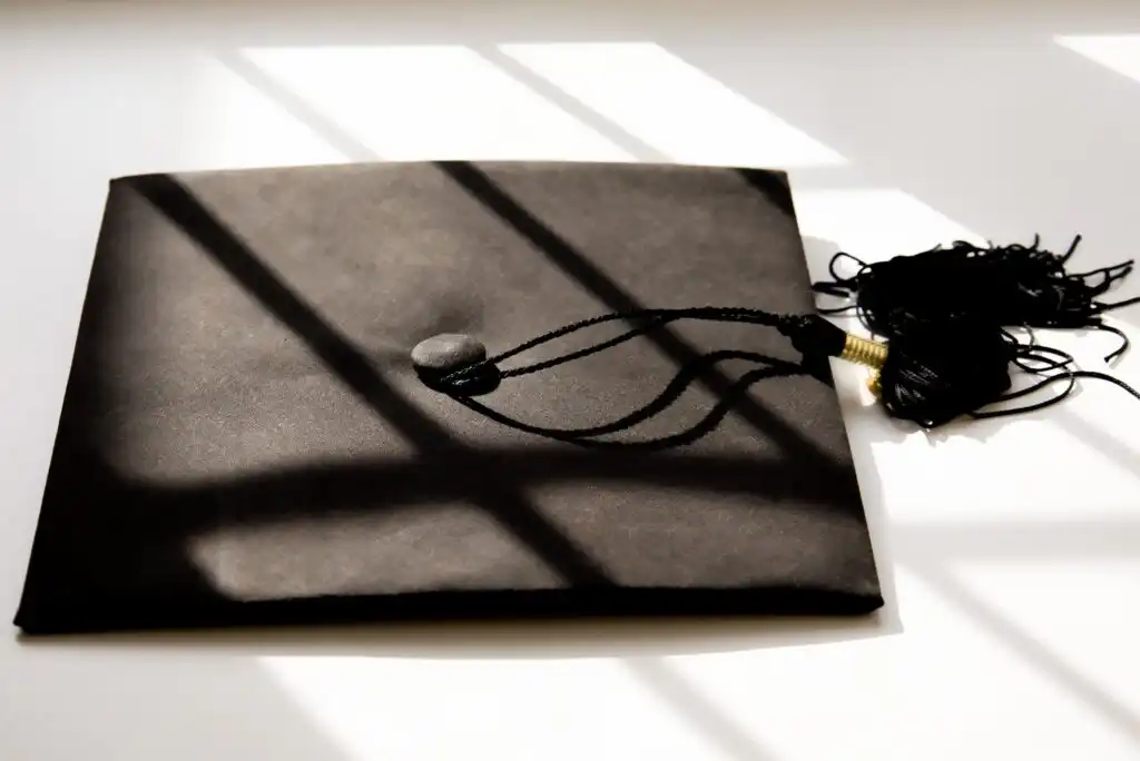 A black graduation cap with a tassel resting on a white surface, with shadows from a window cast across it.