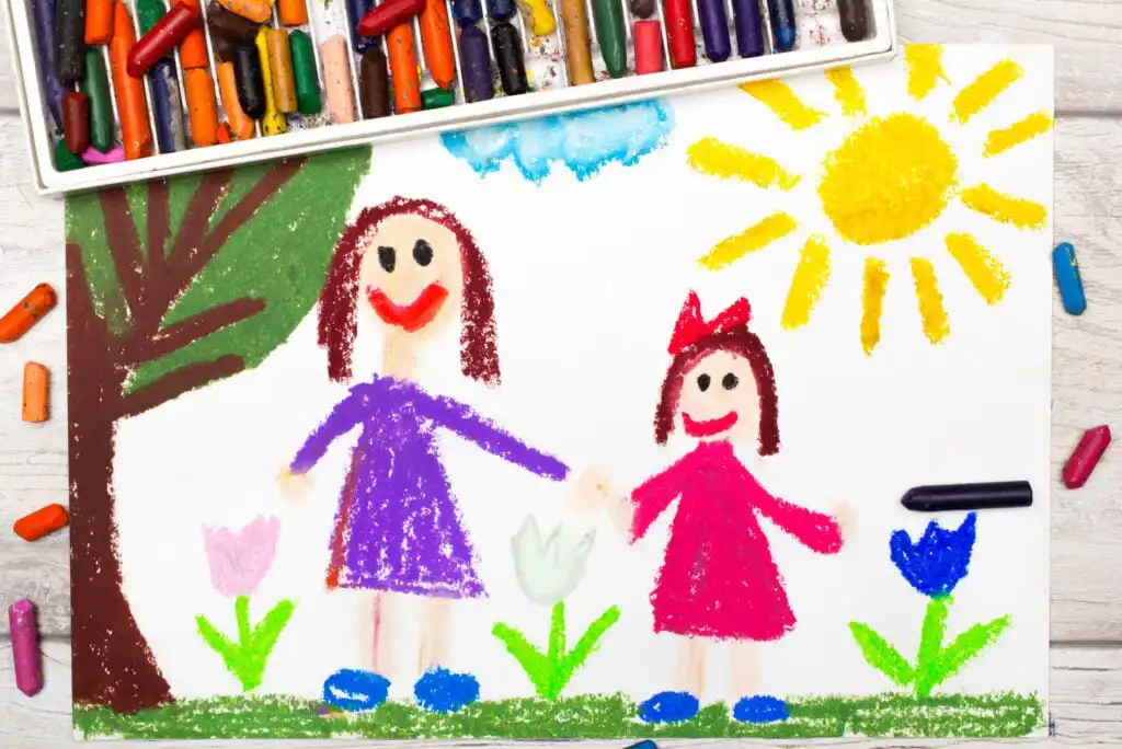 A colorful crayon drawing of two smiling people, one taller and one shorter, holding hands outdoors with flowers, a tree, and a bright yellow sun. Crayons are scattered above the drawing on a wooden surface.