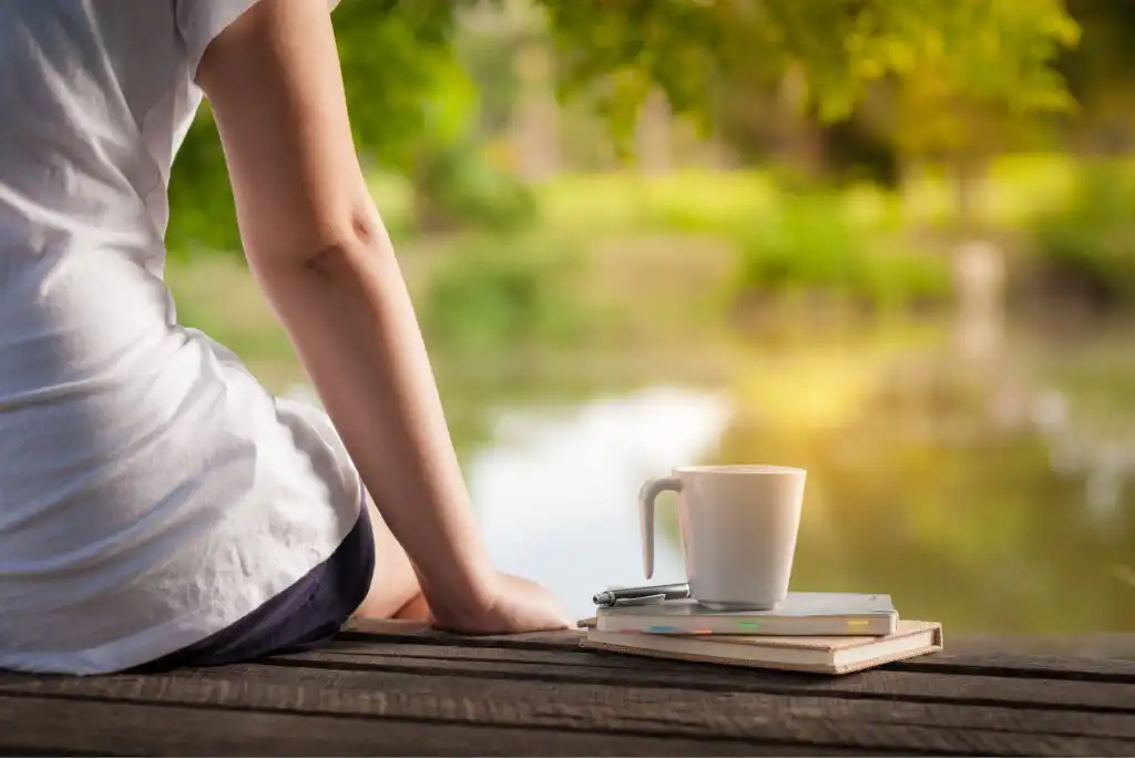 A person sits on a wooden surface near a calm body of water, with a white mug and notebooks beside them. Green trees are reflected in the water, creating a peaceful outdoor scene.