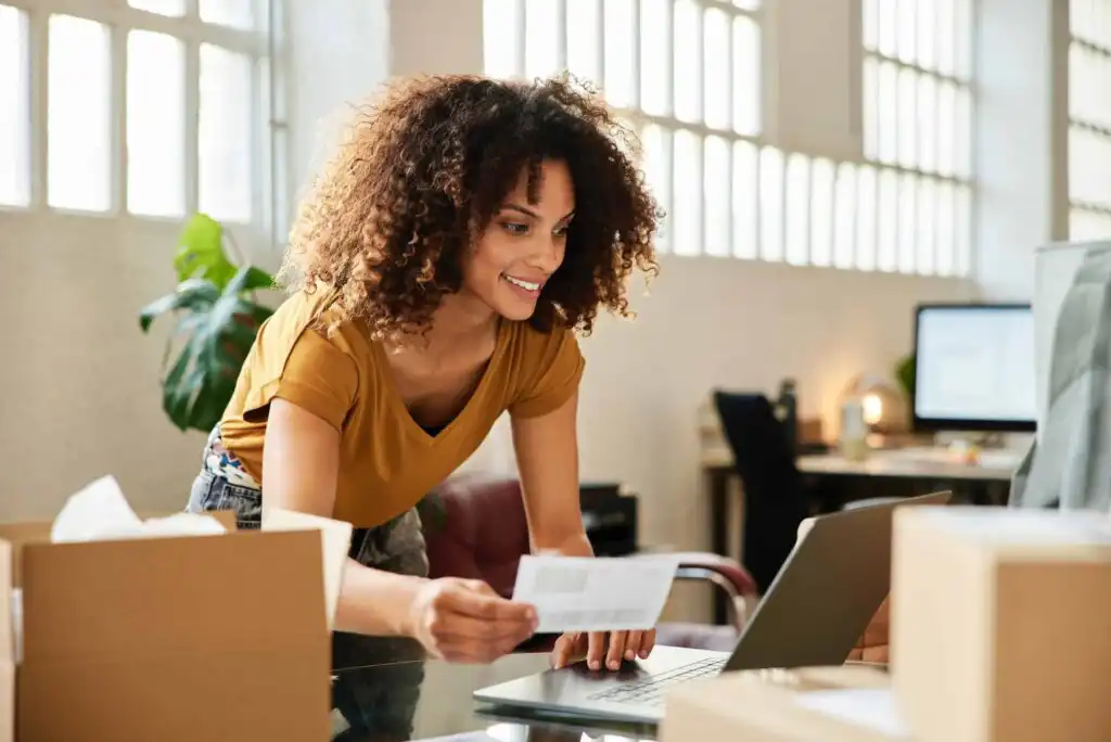 A woman with curly hair smiles while looking at a laptop, holding a piece of paper. She is standing in a bright room with large windows, surrounded by cardboard boxes, suggesting she is working on a small business or shipping orders.