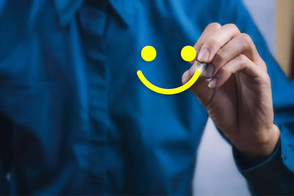A person in a blue shirt draws a bright yellow smiley face on clear glass or a transparent surface with a marker.
