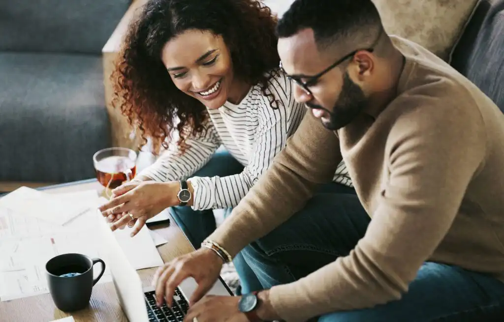 A smiling woman and man sit together at a table, looking at a laptop. Papers, a cup of coffee, and a glass of tea are on the table, suggesting they are working or studying together at home.