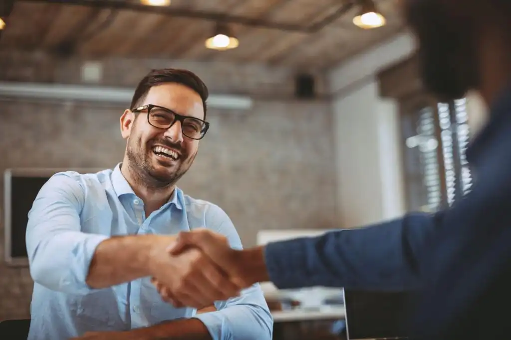 A smiling man in glasses and a light blue shirt shakes hands with another person in an office setting with a laptop on the table. The atmosphere appears friendly and professional.