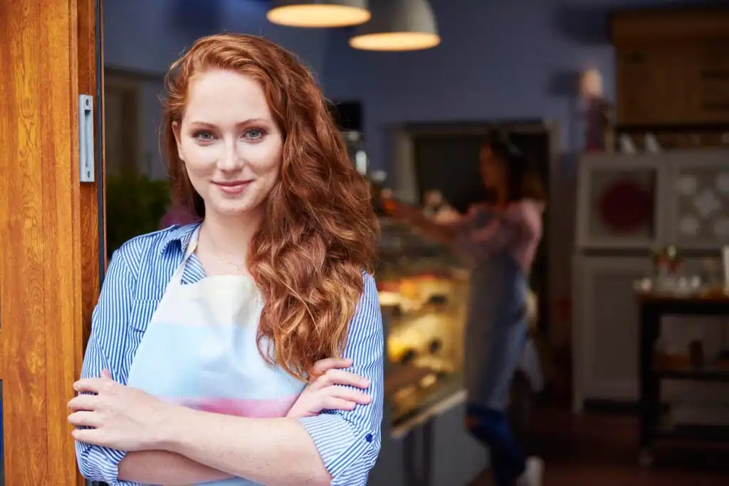 A woman with long red hair wearing a striped shirt and apron stands confidently with folded arms in a bakery doorway. The background shows another person working behind a counter with pastries.