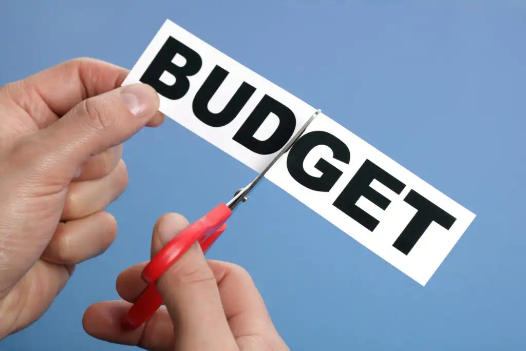 A person uses red scissors to cut a piece of paper with the word BUDGET printed in bold black letters on a blue background.